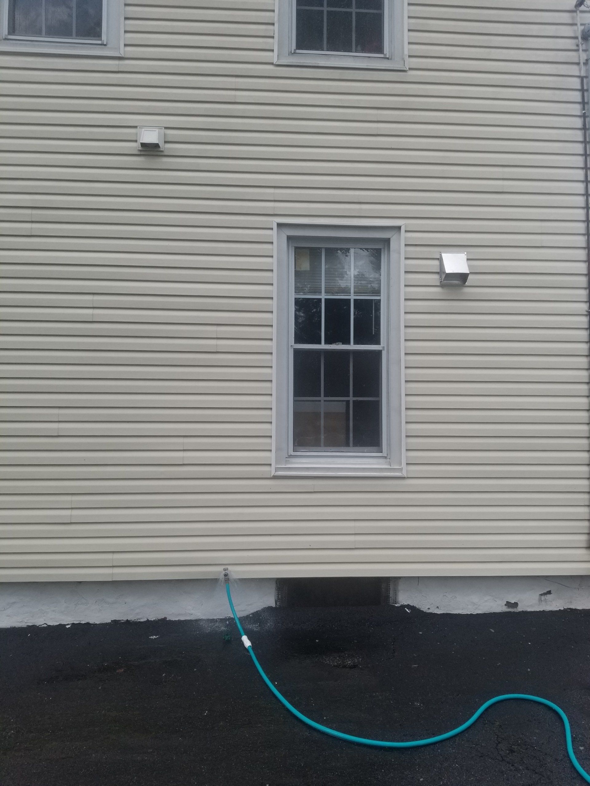 Tan vinyl-sided house exterior with two windows, two vents, and a green garden hose resting on the paved ground.