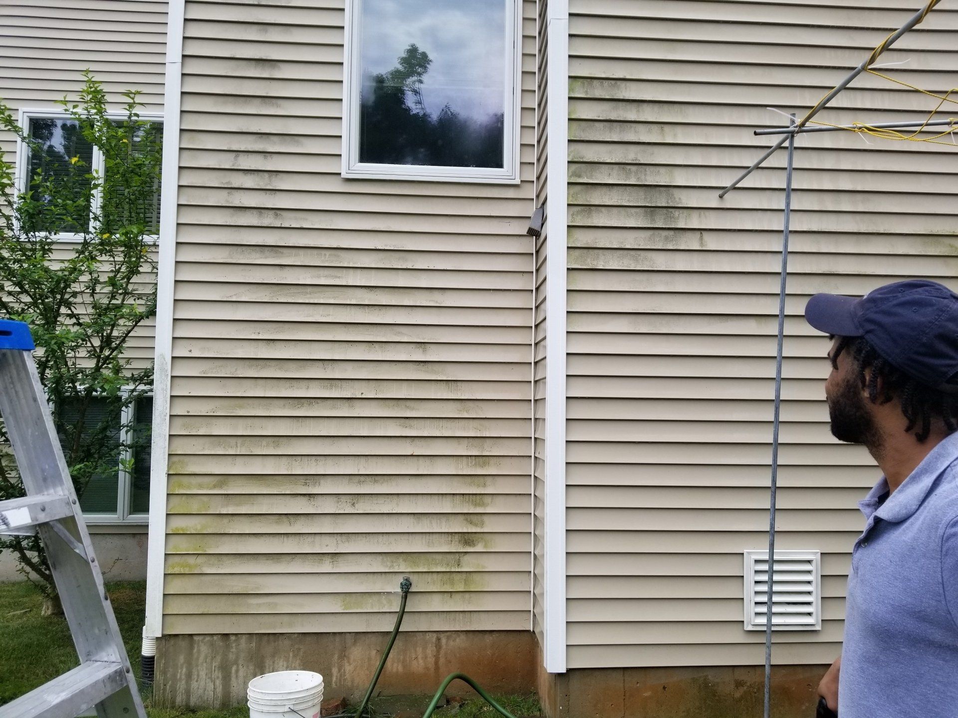 A person in a blue polo and cap stands near a house with dirty beige vinyl siding and an aluminum ladder.