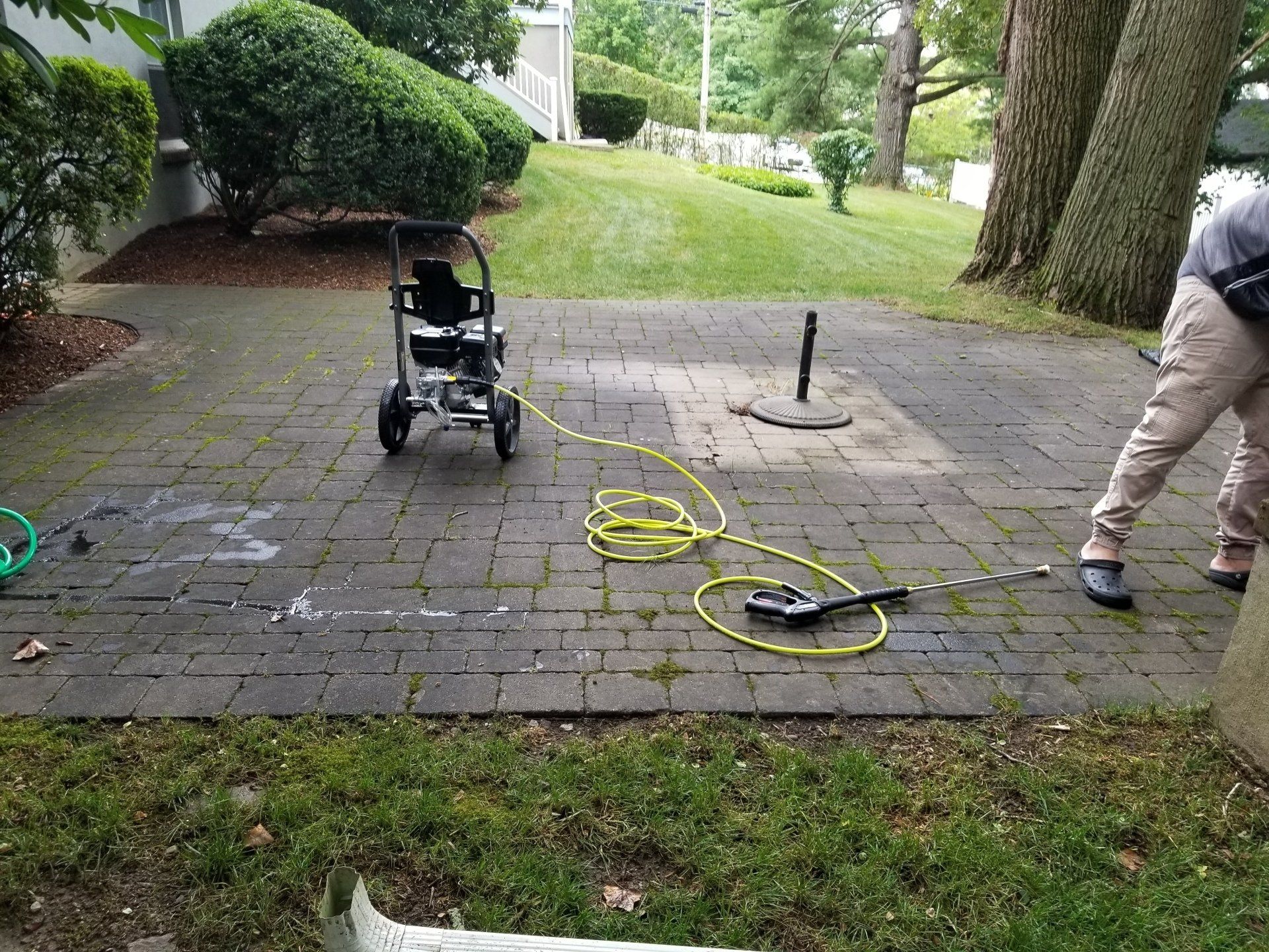 A pressure washer with a coiled yellow hose sits on a paved patio next to a person's legs.