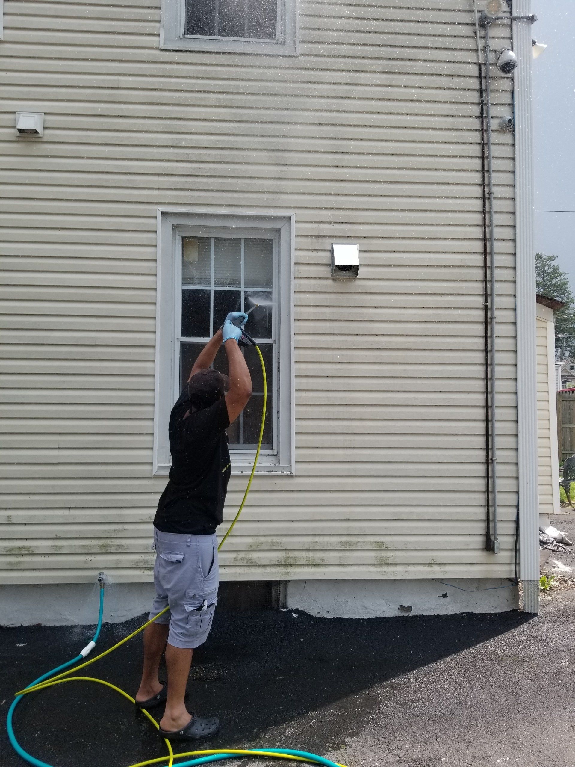 A person in a black shirt and shorts uses a pressure washer to clean the exterior siding of a beige house.