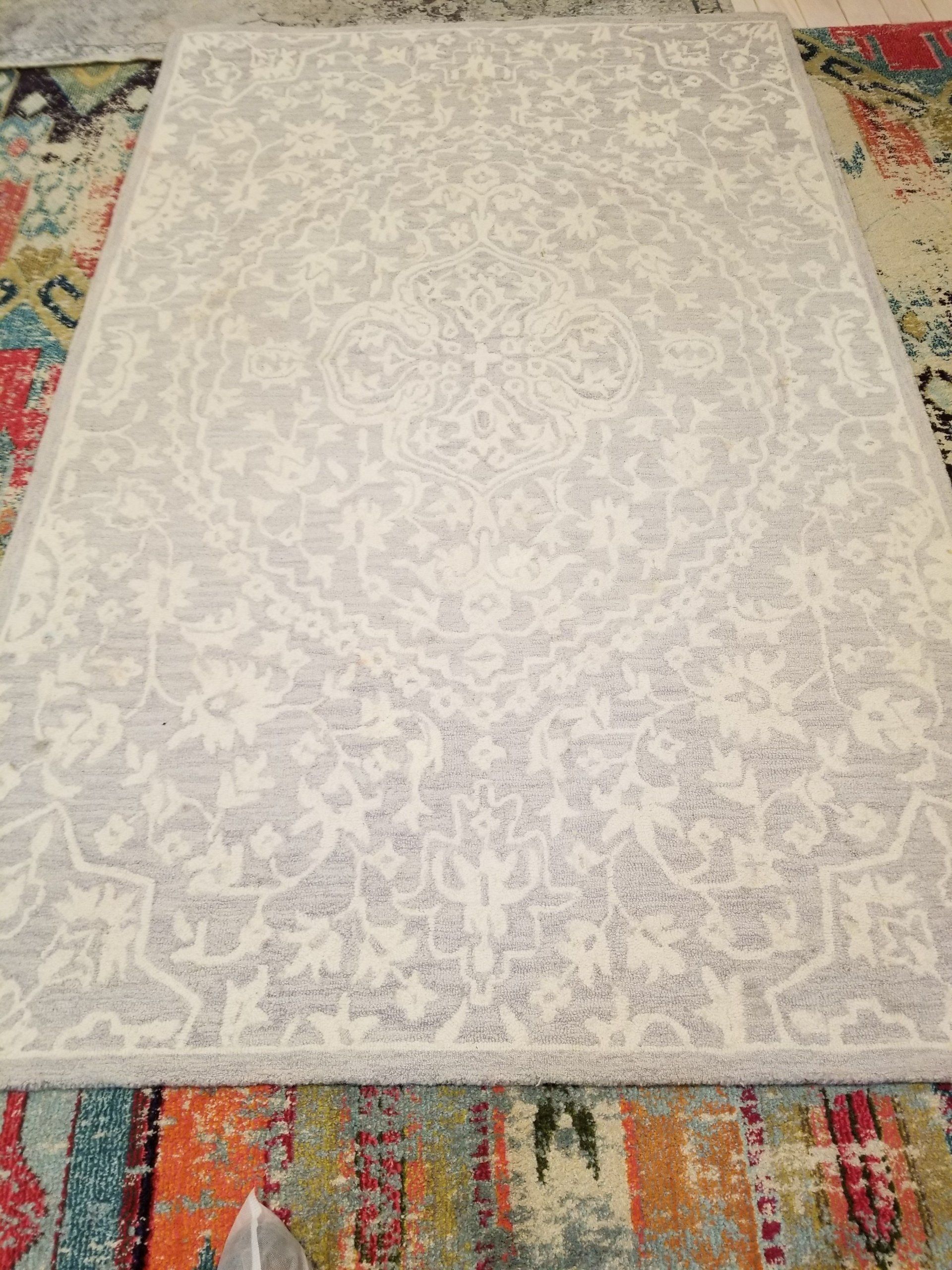 A light grey area rug with a white traditional medallion pattern, layered over a vibrant, multi-colored patterned rug.