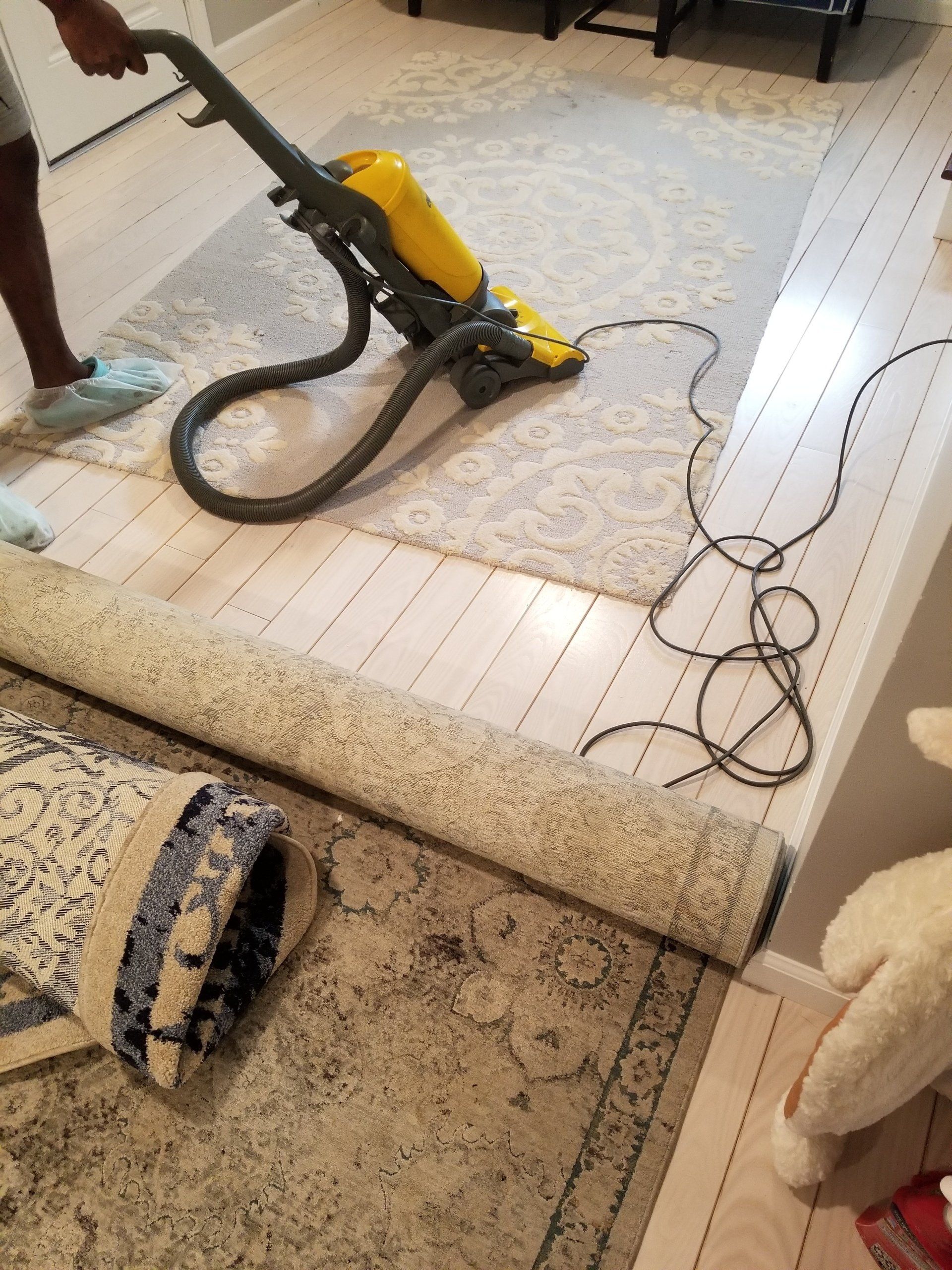 A person uses a yellow vacuum cleaner on a patterned area rug in a room with light-colored wooden floors.