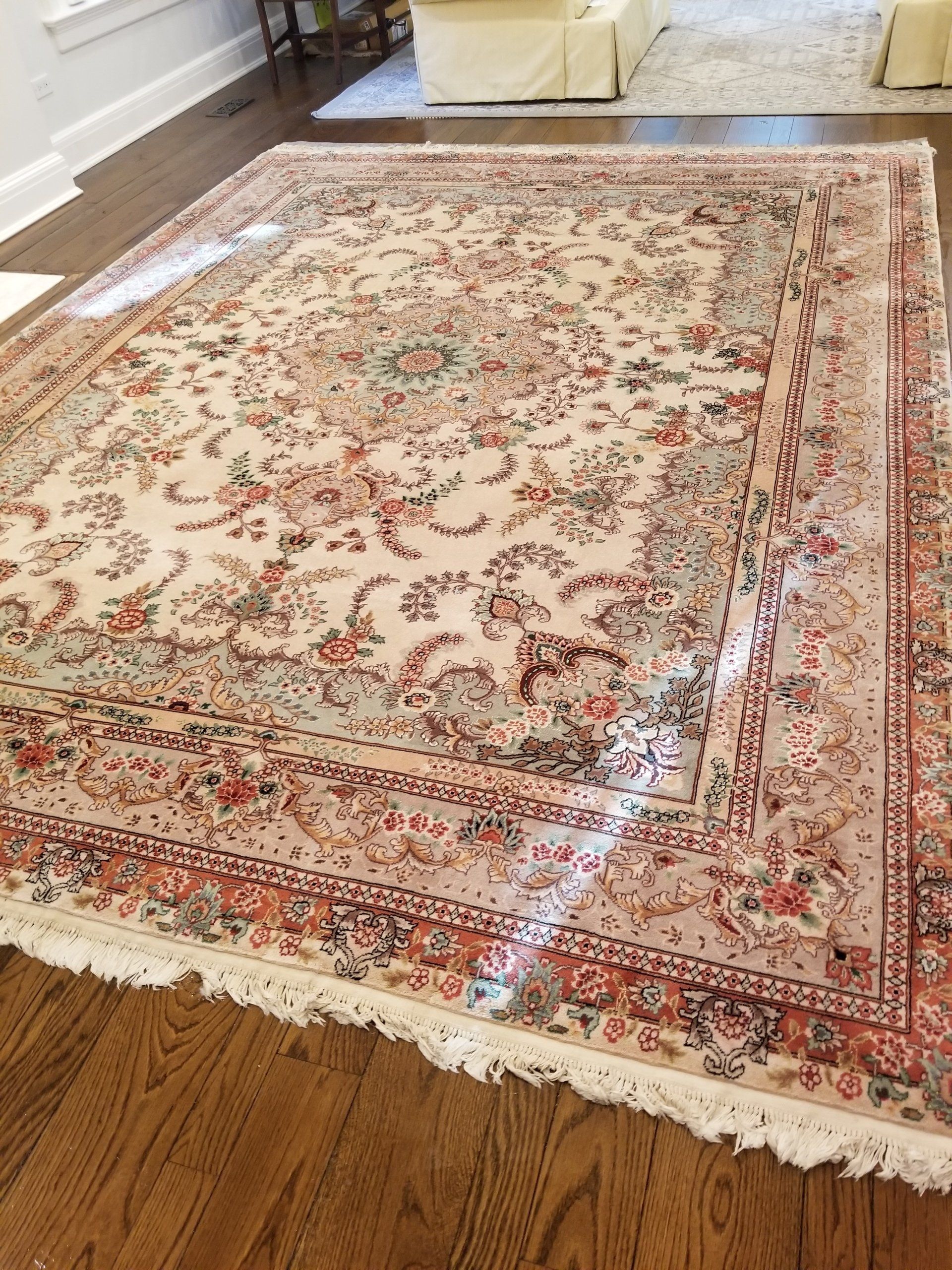 A cream-colored area rug with intricate floral patterns, a central medallion, and fringe on a dark wood floor.