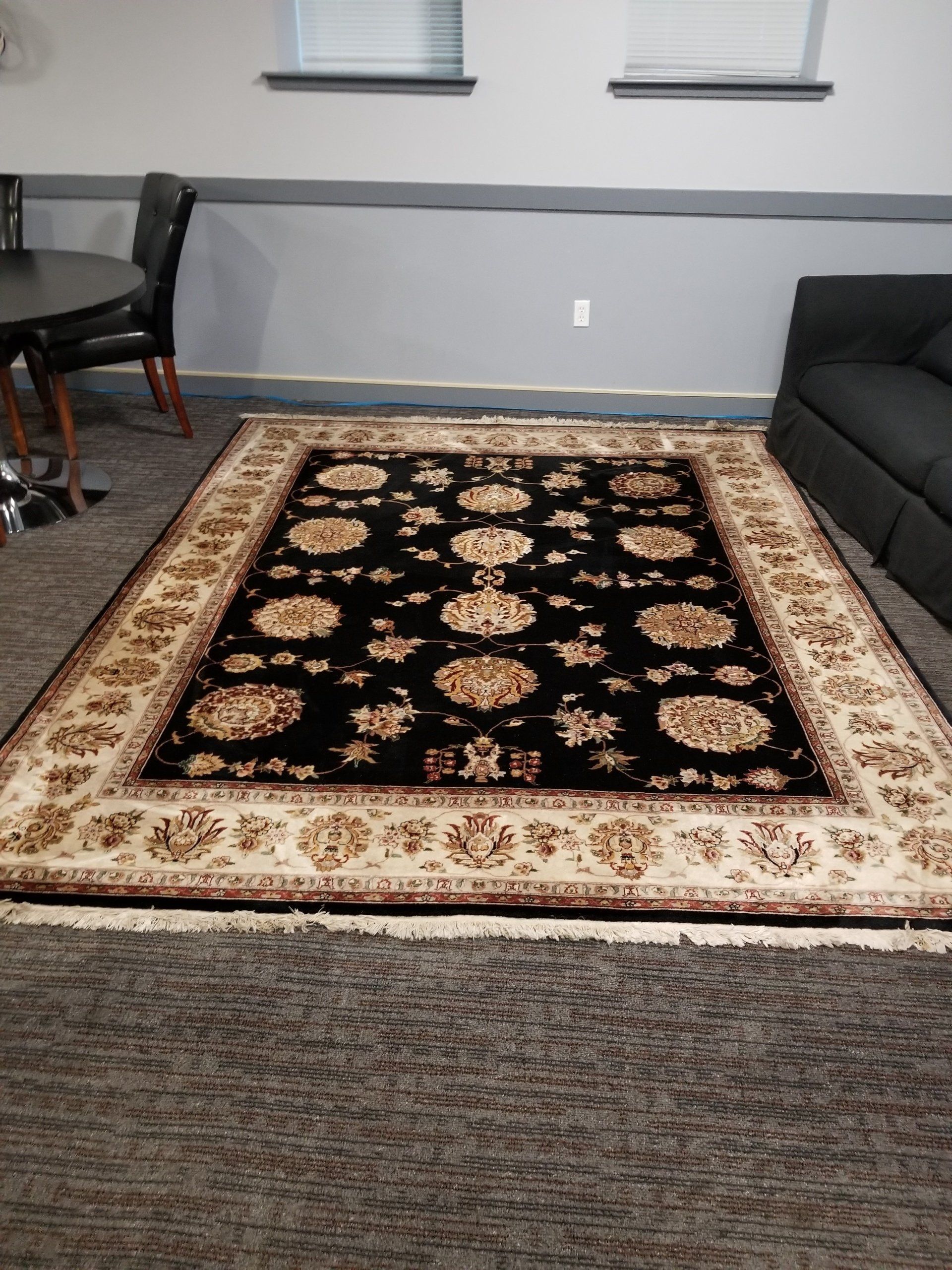A rectangular rug with a black floral center and wide, beige patterned border lies on a gray carpet in a furnished room.