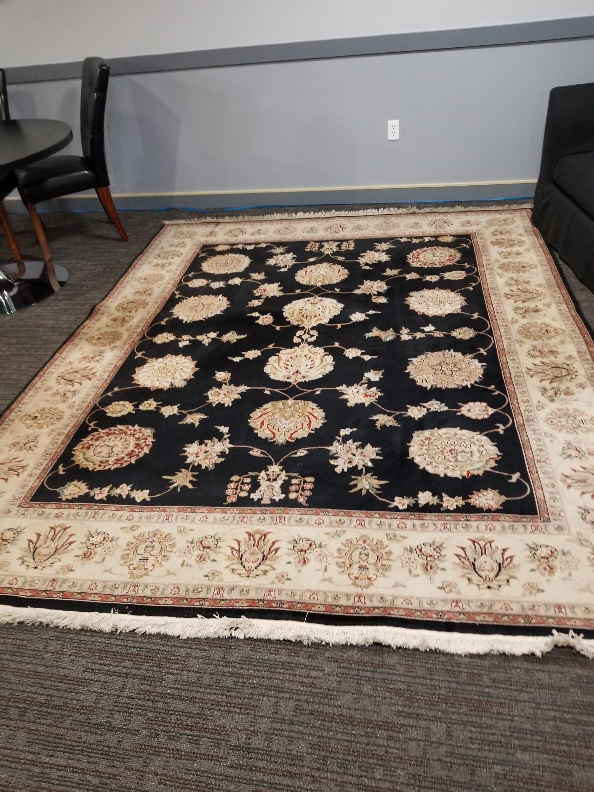 A large rectangular area rug with a black field, ornate cream-colored floral patterns, and a wide, detailed border.