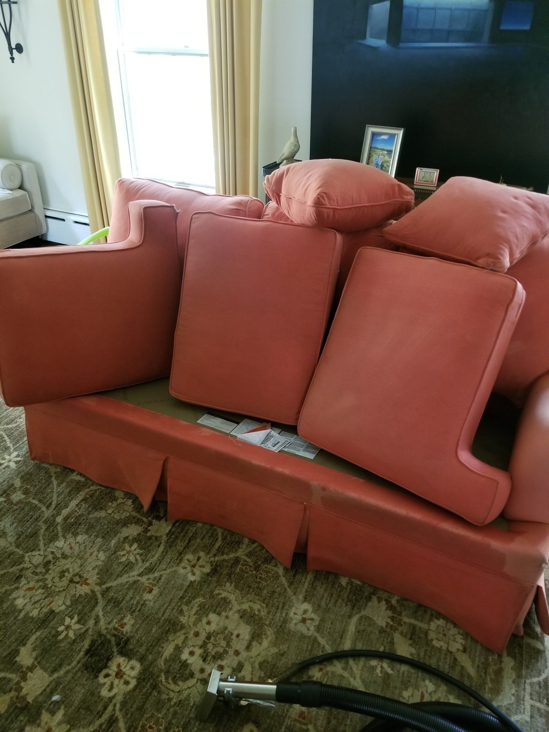A pinkish-coral upholstered sofa with back cushions sits on a patterned rug near a window with floor-length curtains.