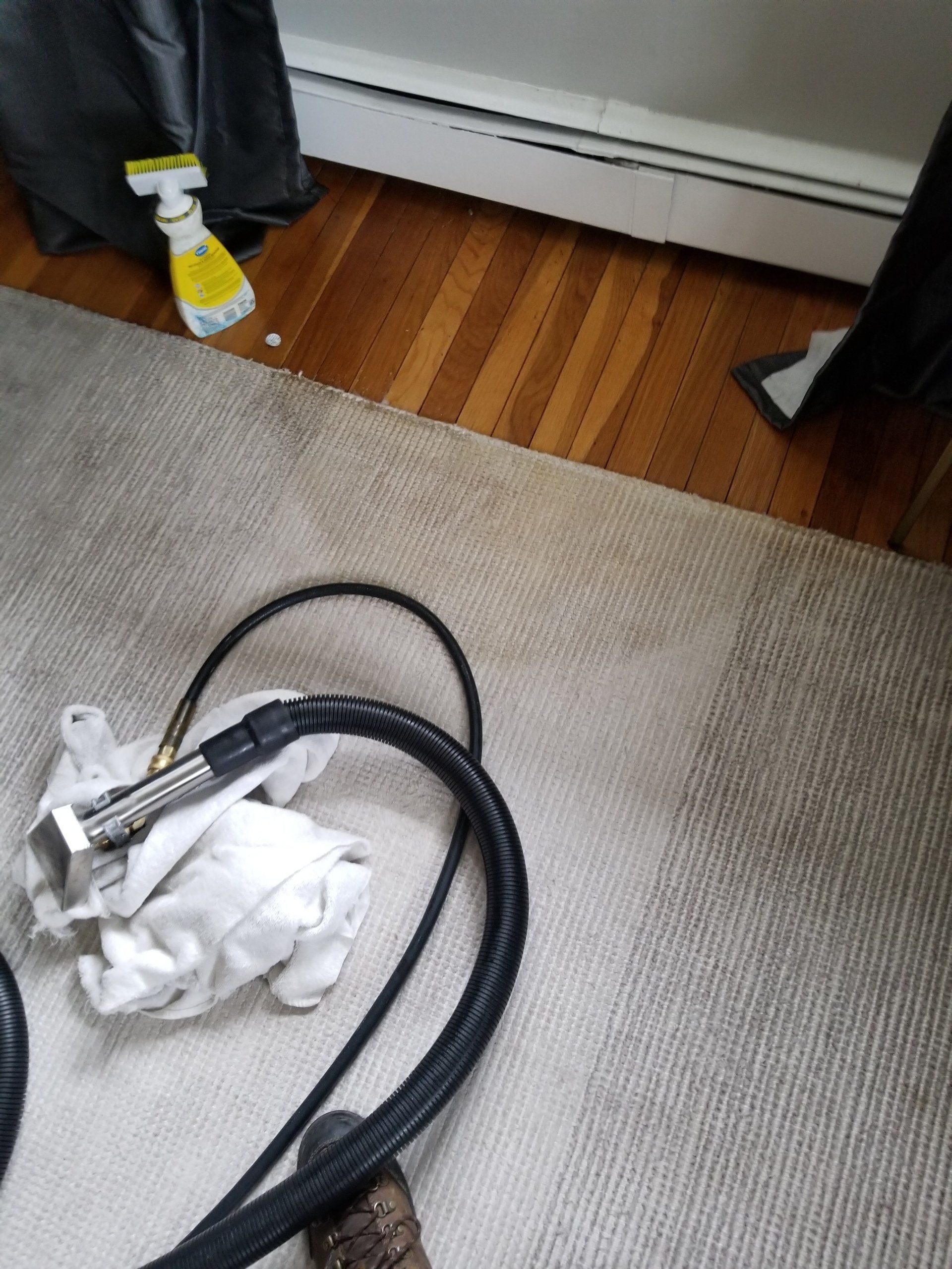A cleaning hose attachment sits on a carpeted floor next to a white spray bottle and a baseboard heater.