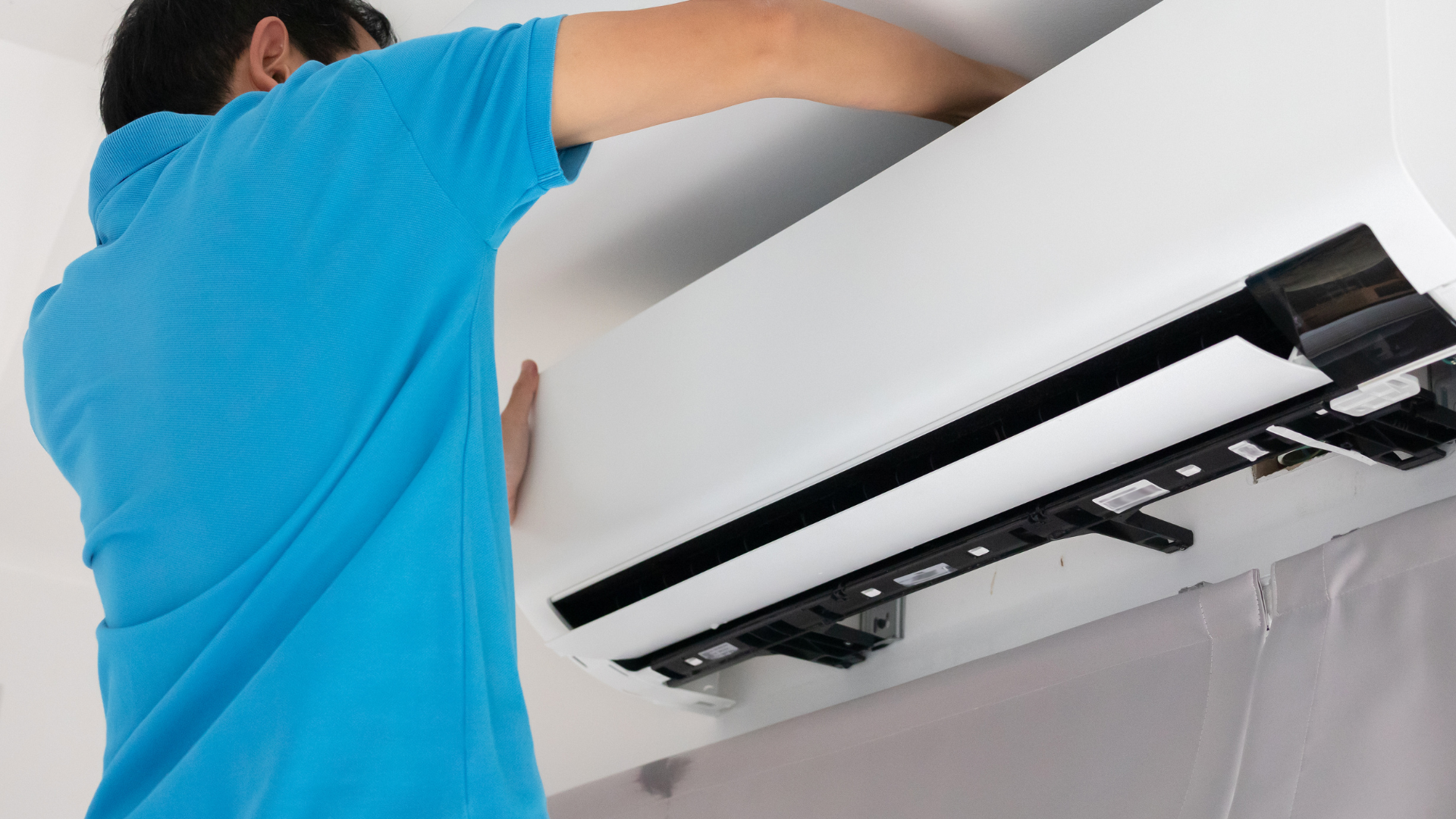 Technician cleaning a wall-mounted air conditioner filter, seen from behind against a white wall.