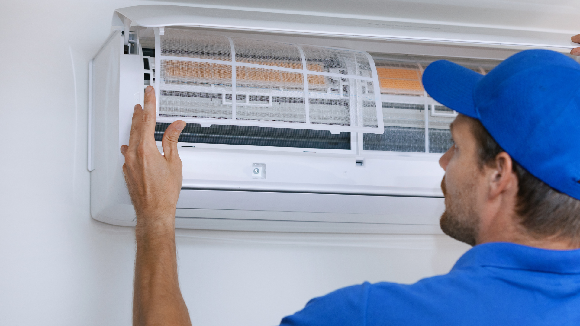 Technician in blue cap cleaning an indoor air conditioner filter on a white wall unit