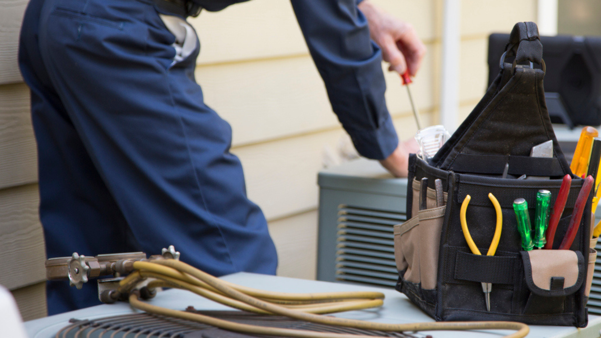 Technician beside HVAC unit with tools bag and hoses on a home exterior.