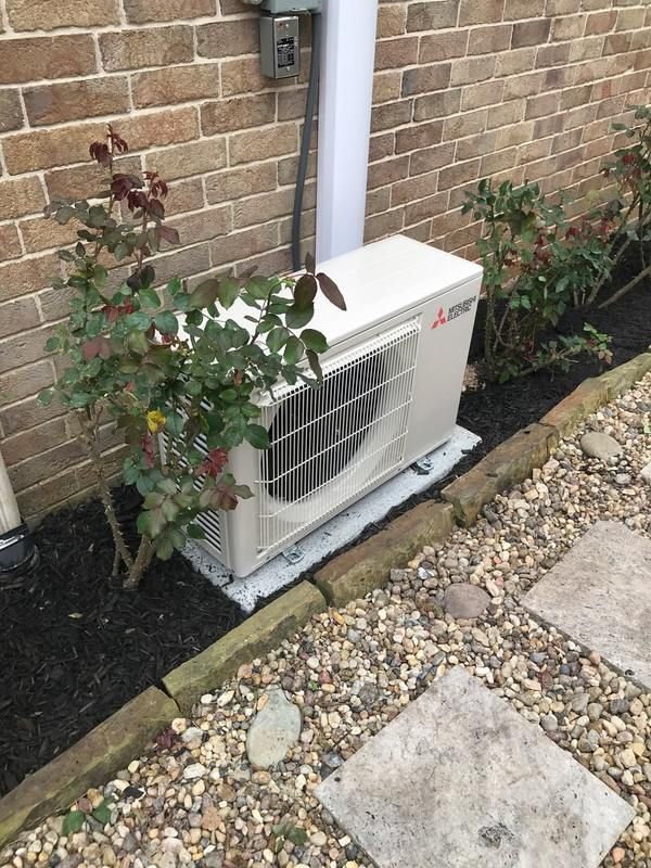 Outdoor air conditioning unit next to brick wall, with bushes and decorative stones.