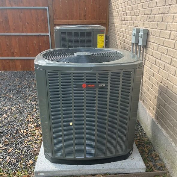 Two Trane air conditioning units, gray, brick wall background, on concrete pads.