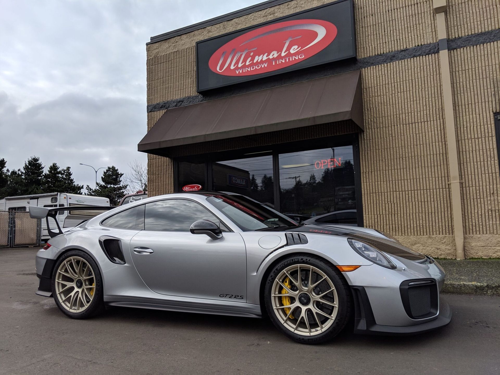 Silver Porsche sports car parked outside a Welcome sign on a rainy day.