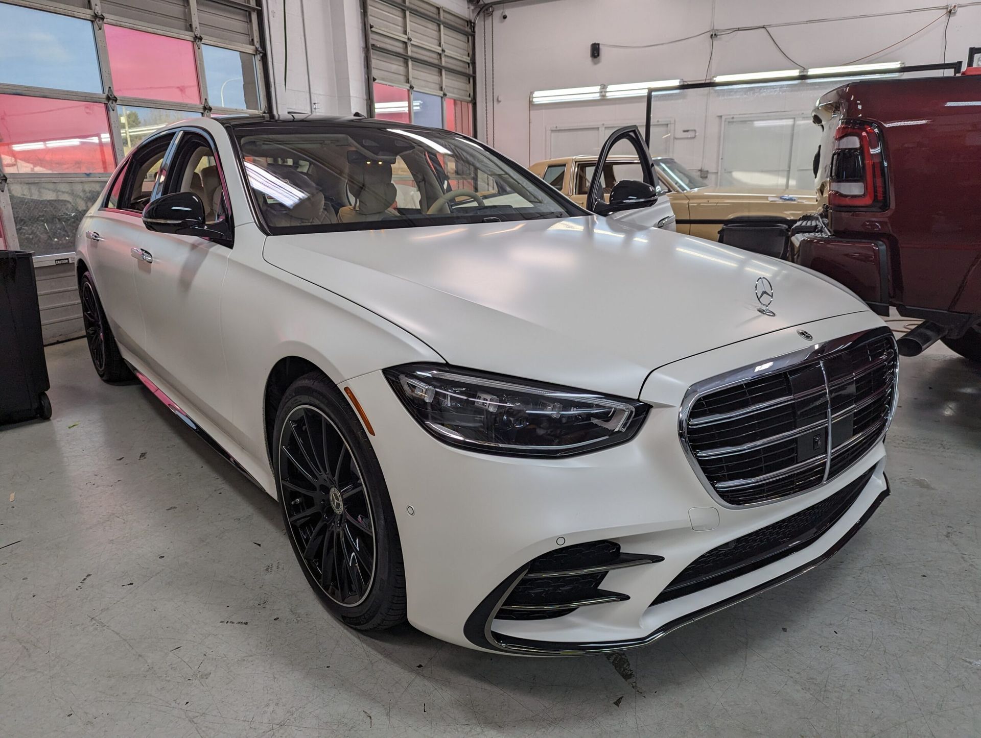 White Mercedes sedan in an auto shop, viewed from the front-left with doors open and a red SUV behind it
