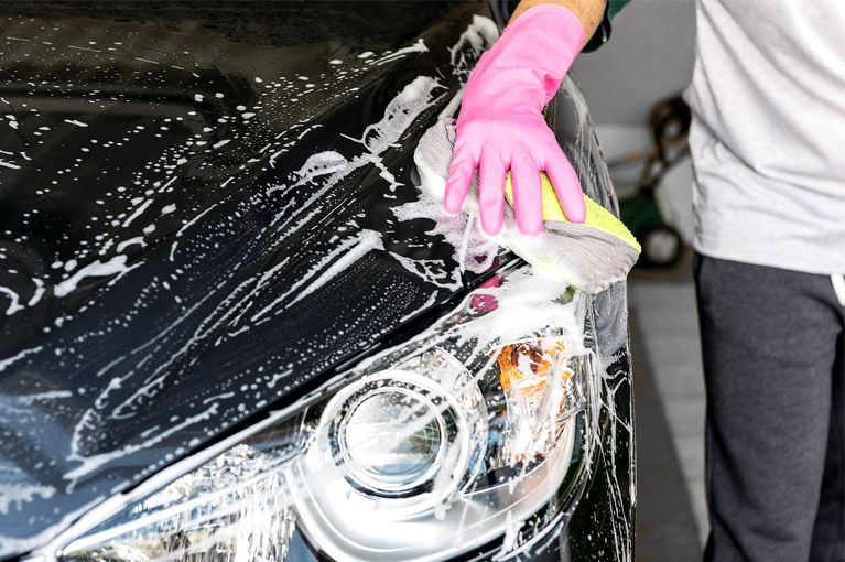 Person washing a black car with soap foam, scrubbing the headlight with a pink glove