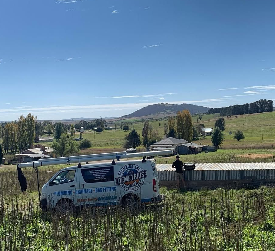 A white van with a ladder parked in a field, a person sits near a structure, a mountain in the background. — Demetrius Projects in Queanbeyan East, NSW