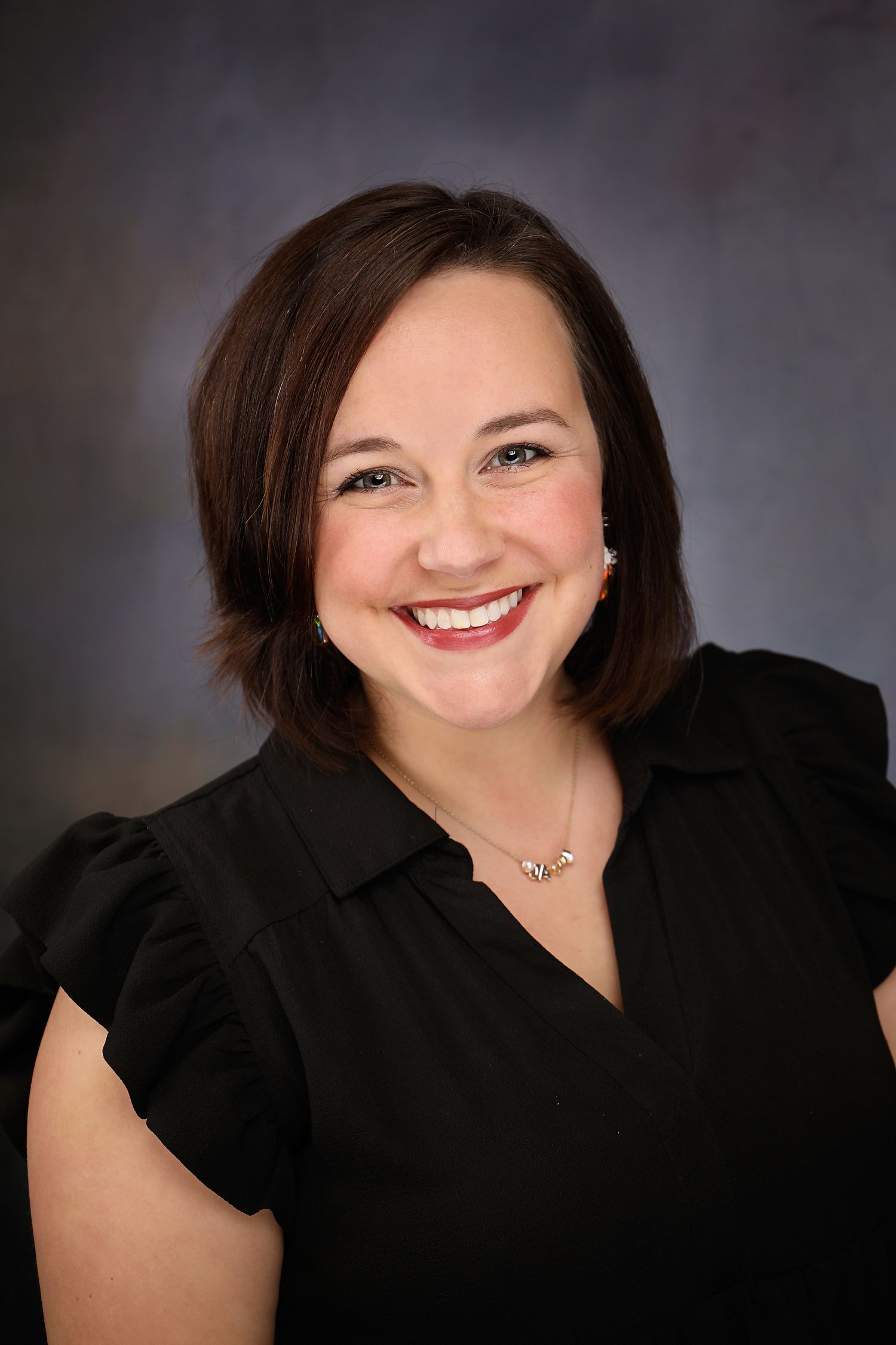 Woman with dark hair, smiling, wearing a black shirt and a necklace, against a blurred background.