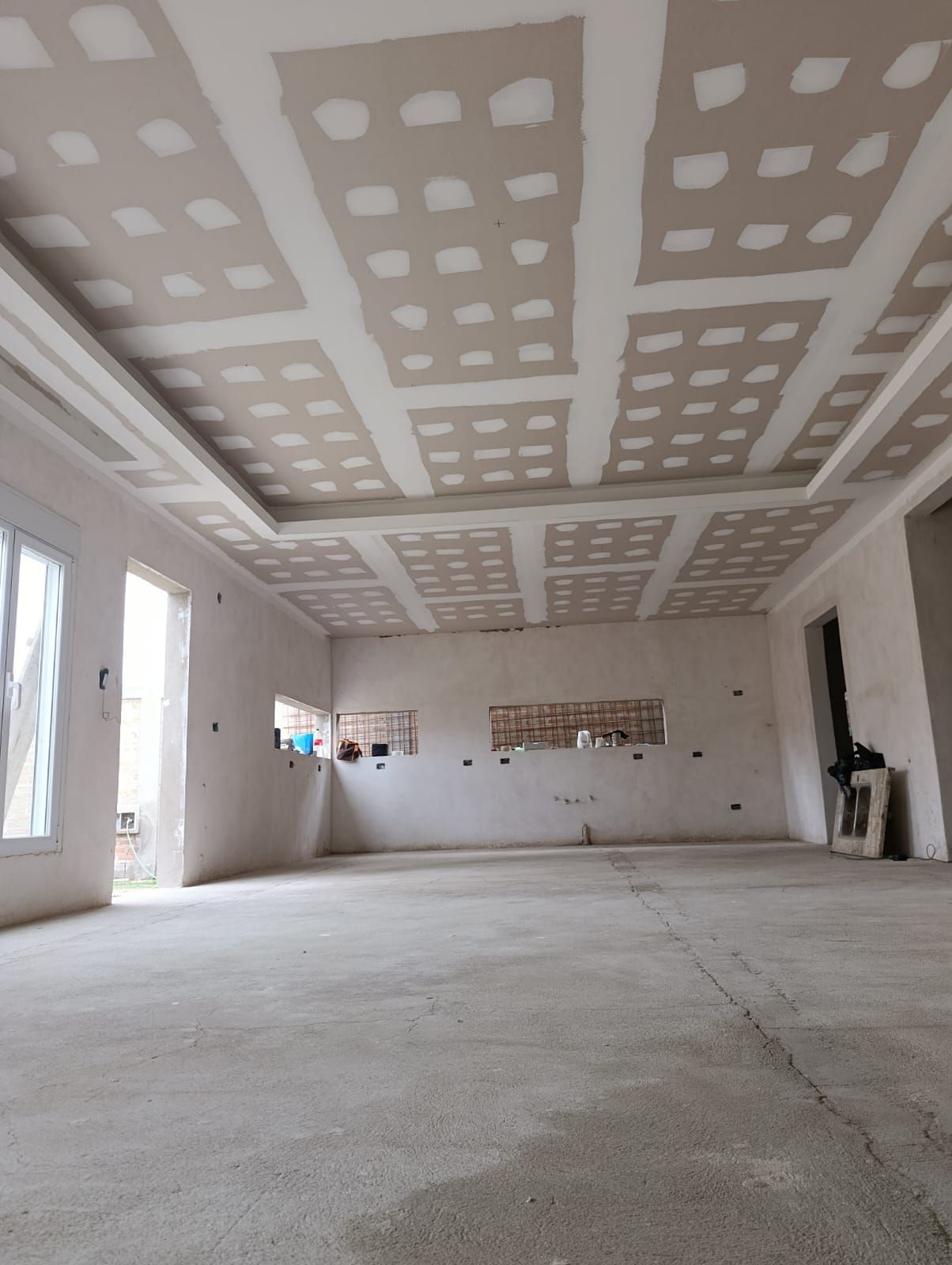 Habitación vacía sin terminar, con parches de pladur en el techo, paredes blancas y suelo de hormigón visto.
