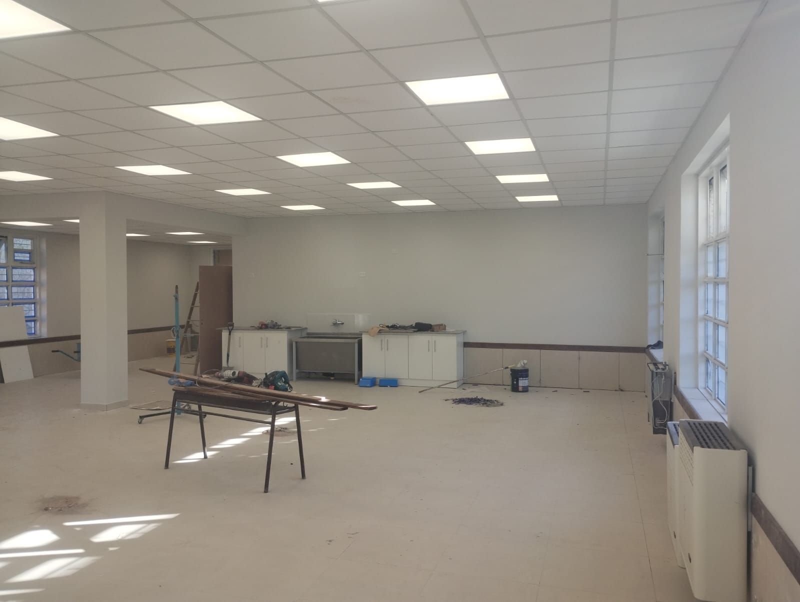 Habitación vacía sin amueblar con techo de baldosas blancas, ventanas luminosas y materiales de construcción en el suelo.