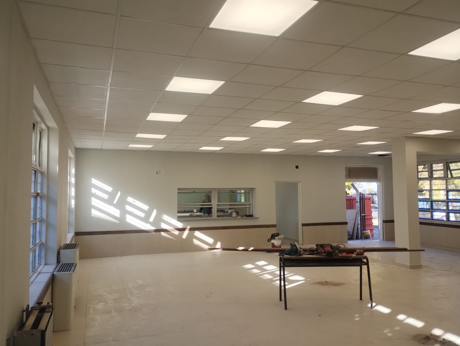 Habitación vacía con paredes blancas, luces fluorescentes y luz solar que entra por las ventanas e ilumina el suelo.