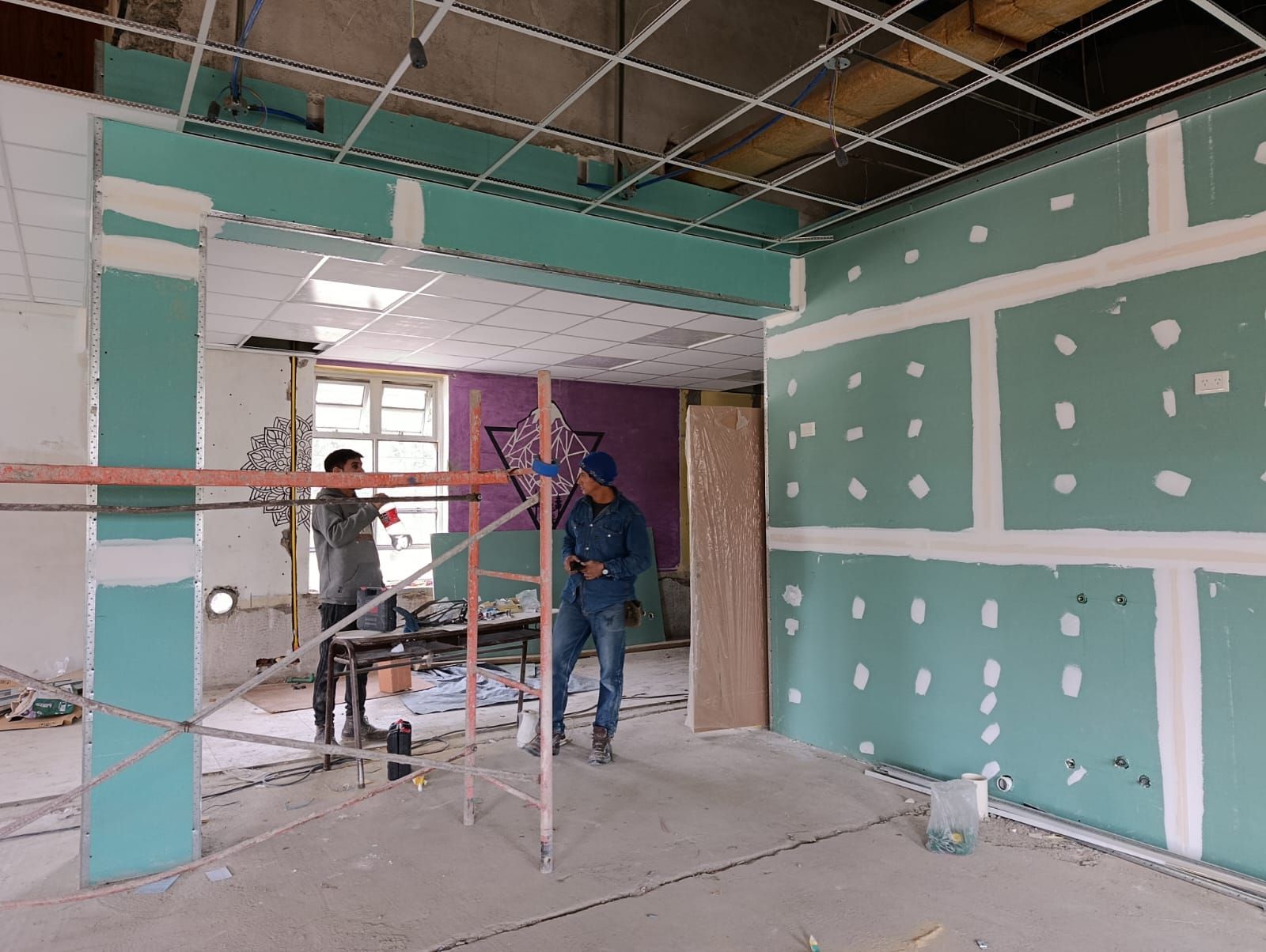 Dos trabajadores en una habitación parcialmente terminada con paneles de yeso, andamios y vigas de techo a la vista.