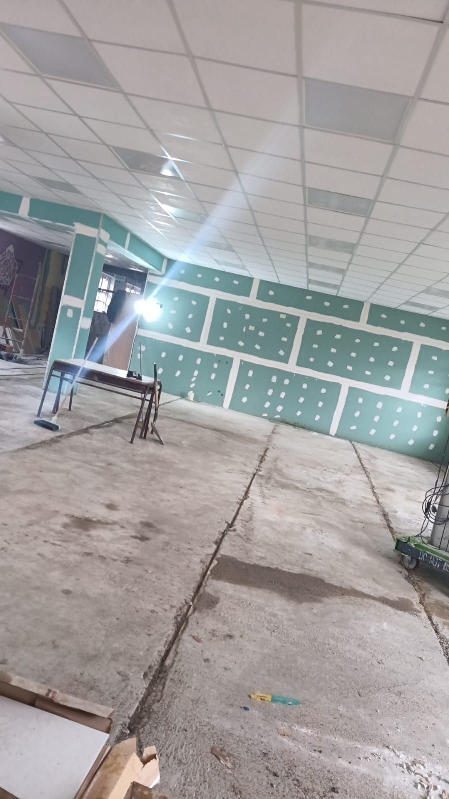 Pasillo interior con suelo de hormigón fresco, paneles de pared verdes y una sola silla bajo un techo de baldosas.