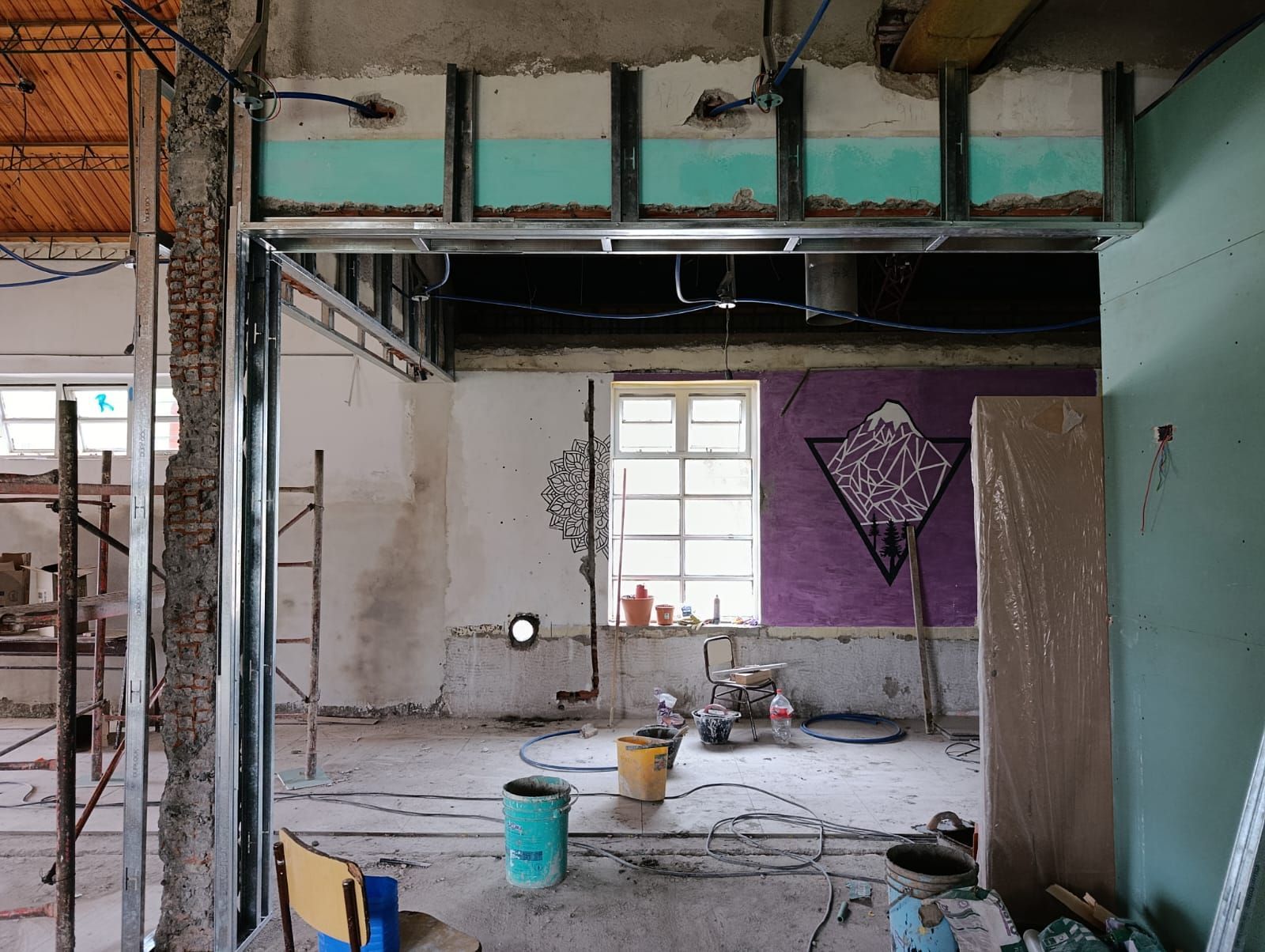Habitación parcialmente renovada con estructura de madera a la vista, escalera, herramientas y una pared morada con ventana.