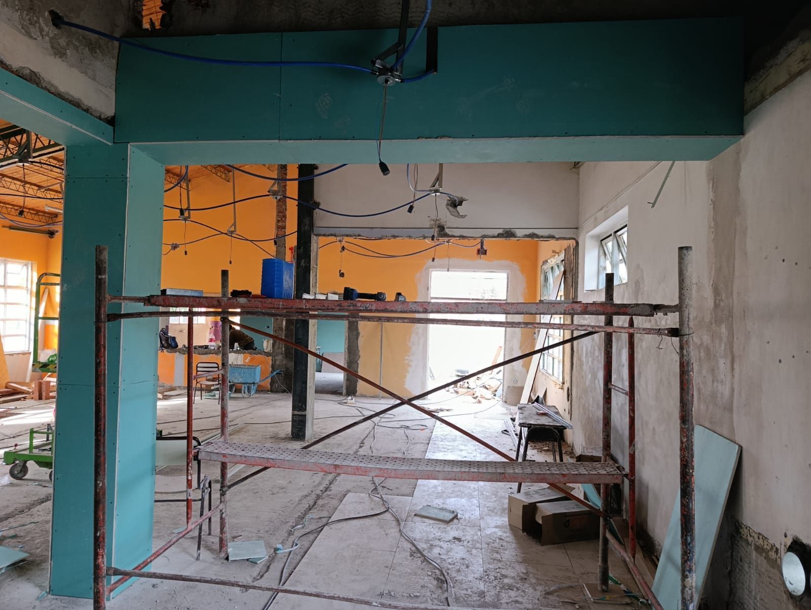 Habitación interior en obras con andamios, vigas color turquesa, paredes naranjas y cableado a la vista.