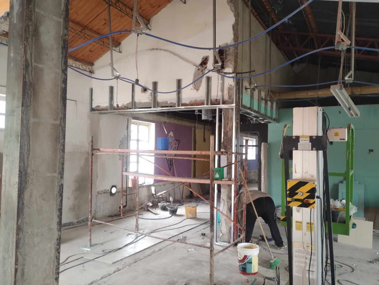 Obra en construcción con andamios, paredes expuestas, cables colgando y un trabajador pintando cerca de una puerta verde.
