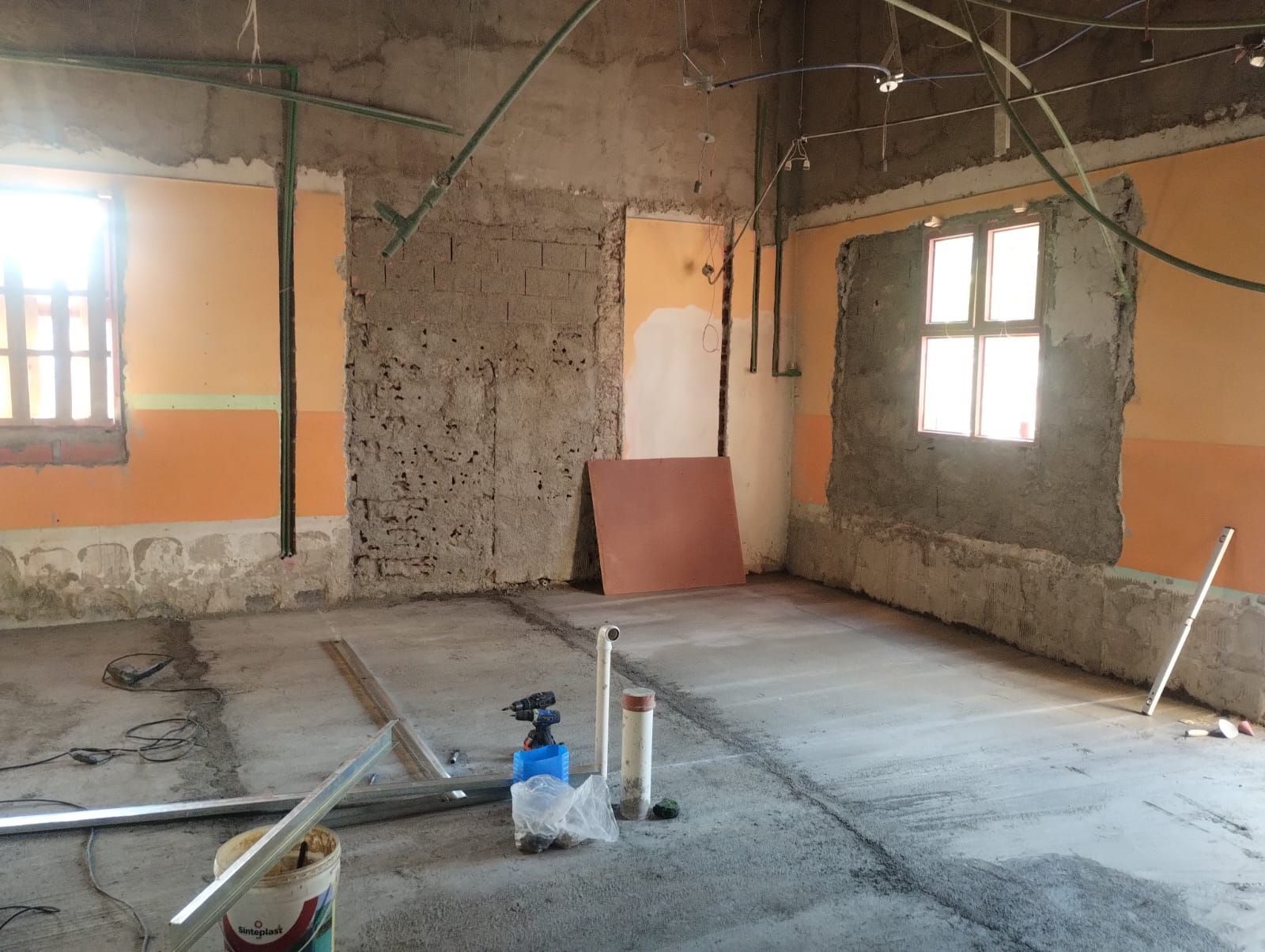 Habitación sin terminar con paredes enlucidas, dos ventanas y materiales de construcción sobre un suelo de hormigón.