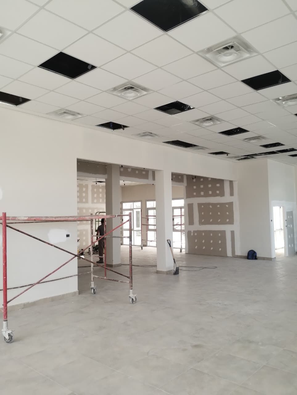 Interior de oficina vacío en obras de renovación con andamios, techos de baldosas a la vista y grandes ventanales.
