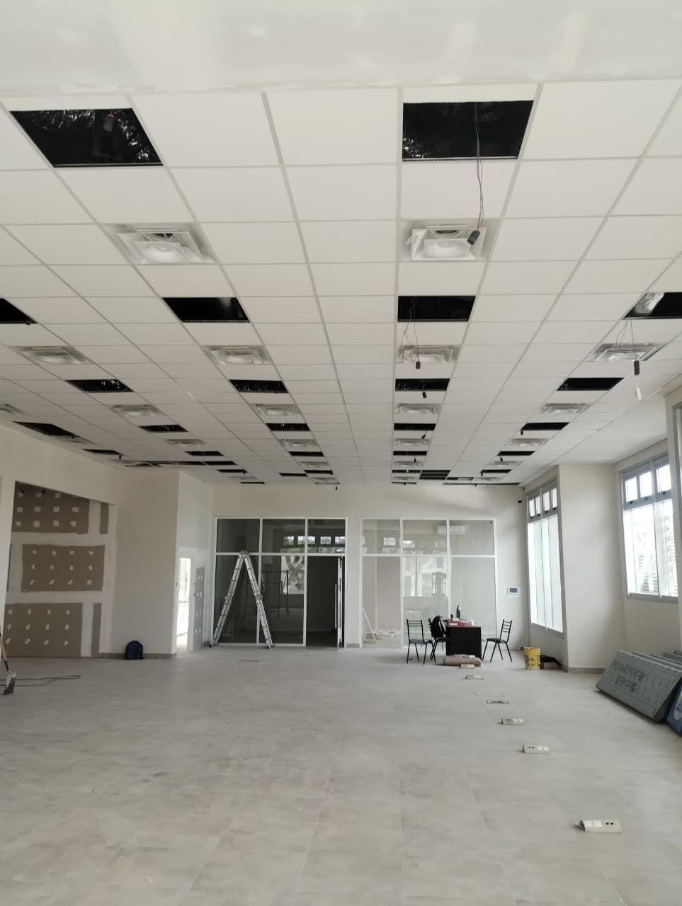 Interior de oficina vacío y sin terminar, con falso techo, ventanas y materiales de construcción en el suelo.