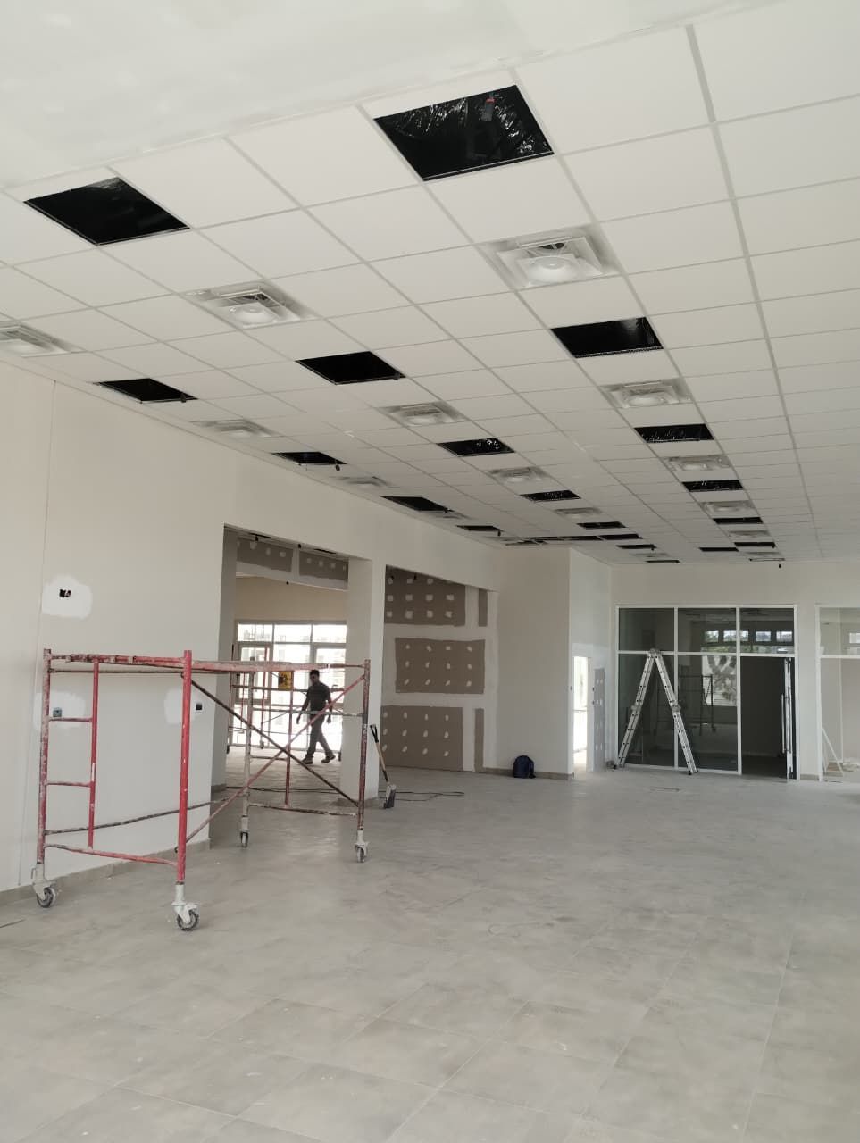 Habitación vacía sin terminar con techo blanco, paneles de rejilla a la vista, andamios y suelo de hormigón.