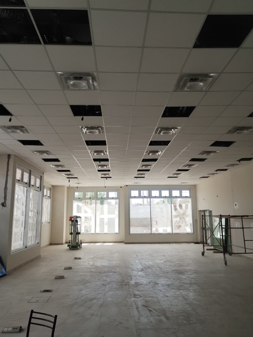 Interior de oficina vacío con falso techo dañado, escombros esparcidos y grandes ventanas en el extremo opuesto.