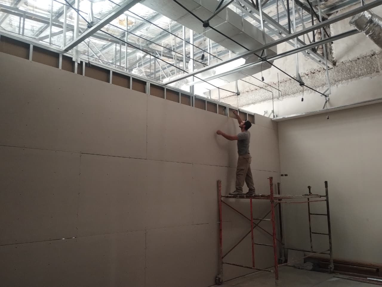 Un obrero pinta en un andamio una gran pared interior bajo un techo con estructura de acero.