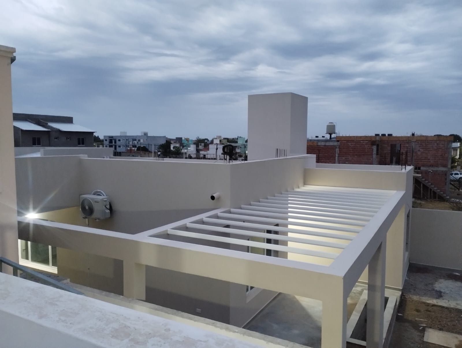Terraza en la azotea con pérgola blanca y cielo nublado sobre edificios de poca altura.