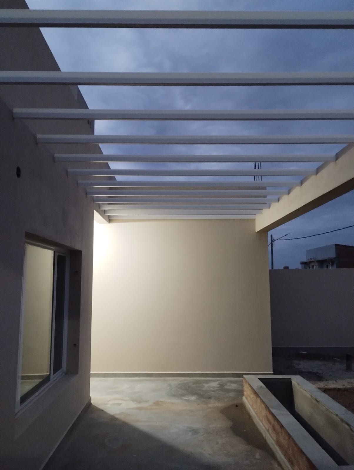 Patio moderno en construcción con vigas blancas, paredes grises y suelo de hormigón al atardecer.