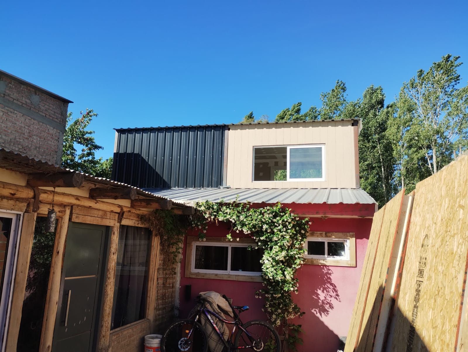Patio trasero con una casita, un cobertizo rojo y una bicicleta bajo un cielo azul.