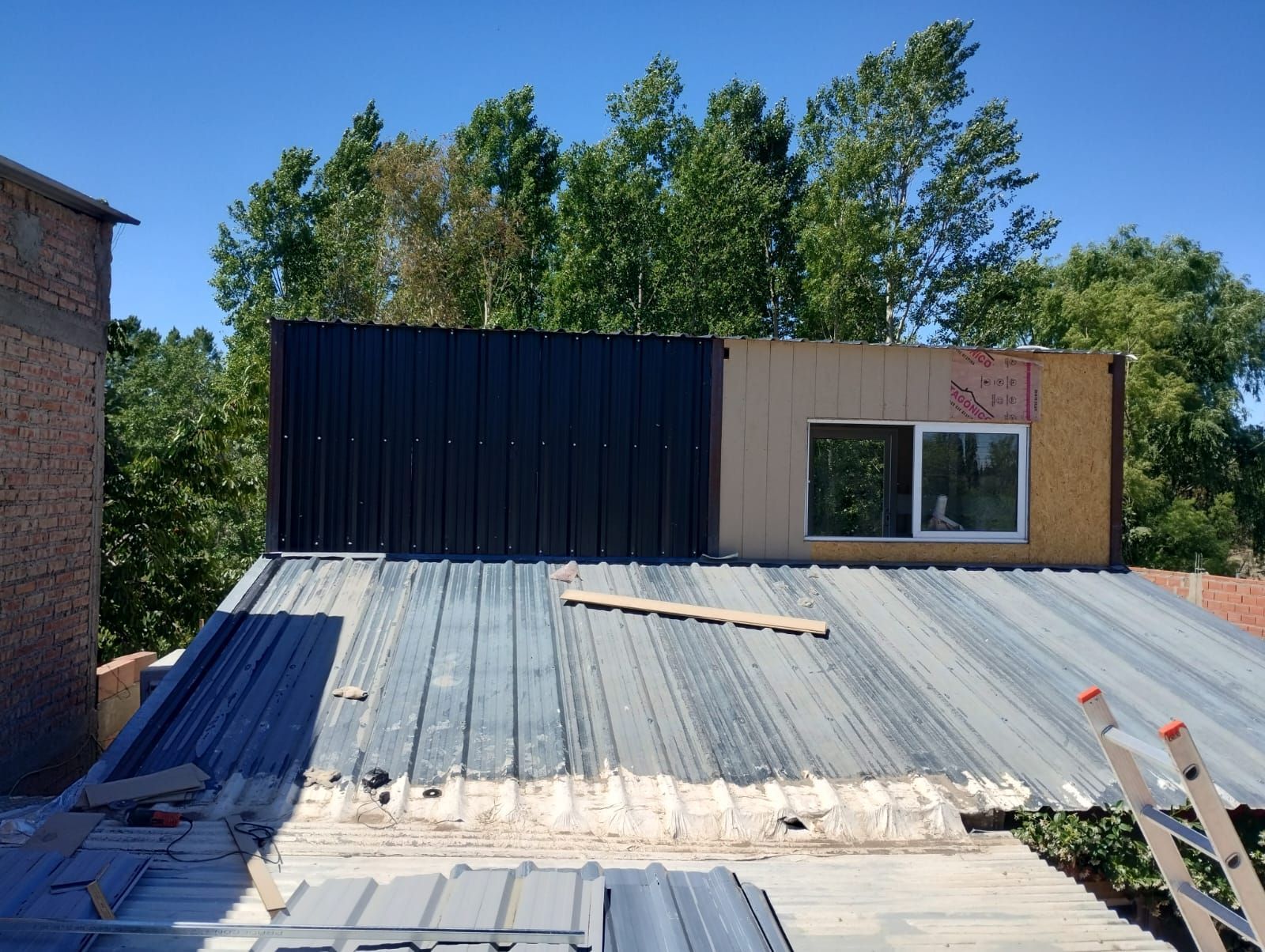 Azotea con paneles de metal corrugado, una pared de madera y negro, y una ventana bajo un cielo azul.