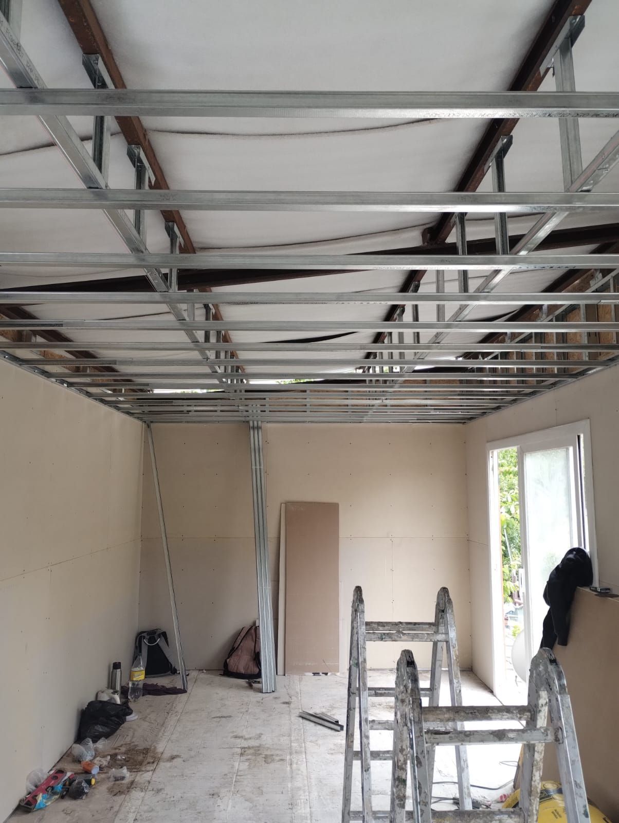 Habitación parcialmente reformada con vigas de techo a la vista, escalera y puerta abierta cerca de una ventana.