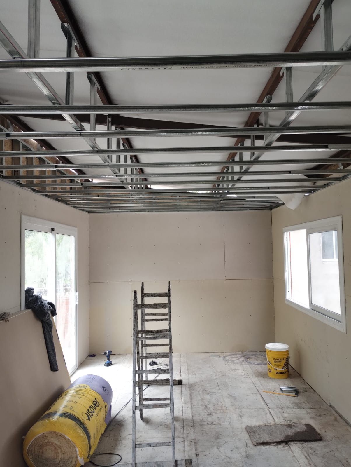 Habitación en obras con estructura de techo a la vista, escalera, paneles de yeso y materiales de construcción en el suelo.