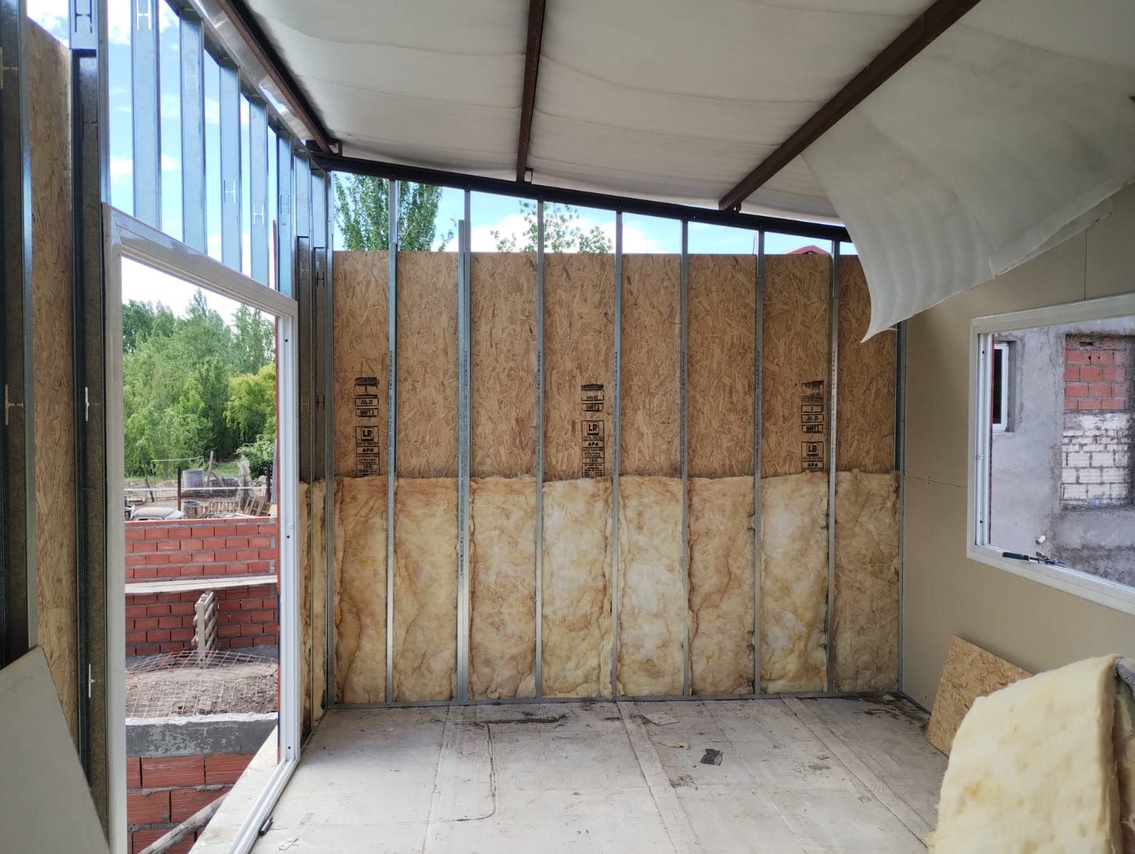 Habitación sin terminar con aislamiento a la vista, estructura metálica y grandes ventanales en construcción.