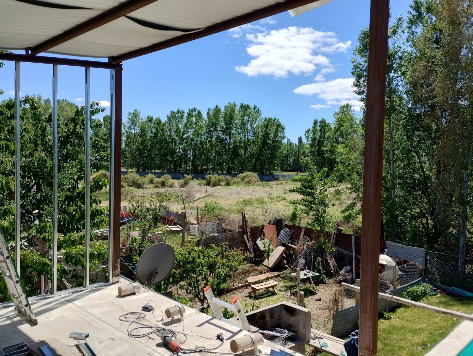 Patio con vistas a un jardín con árboles, cielo azul y escombros de construcción dispersos.