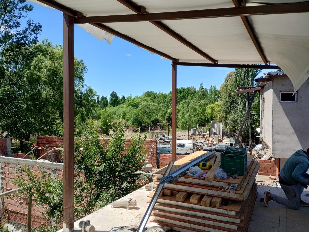 Patio cubierto en construcción junto a un jardín arbolado, con un trabajador agachado a la derecha.