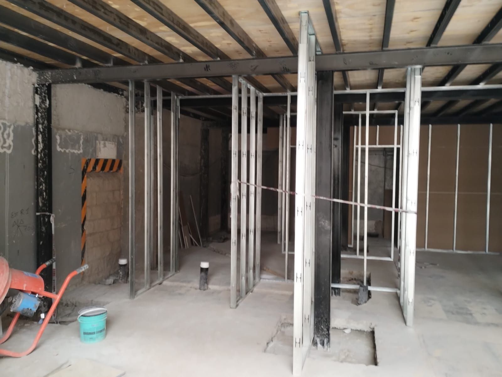 Interior sin terminar con vigas de pared y de techo expuestas, y herramientas de construcción sobre un suelo polvoriento.