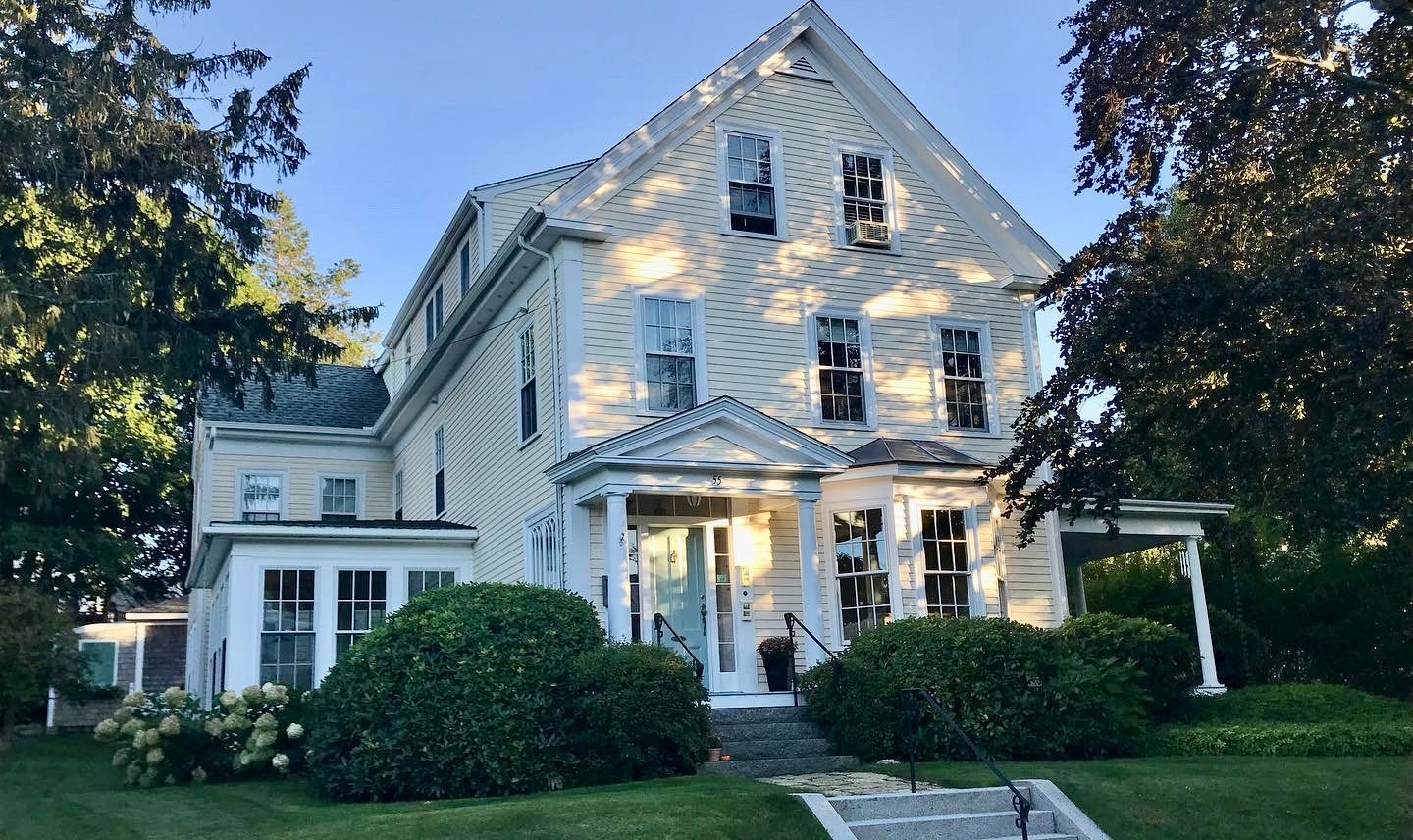 A large white house with a large porch is surrounded by trees and bushes.