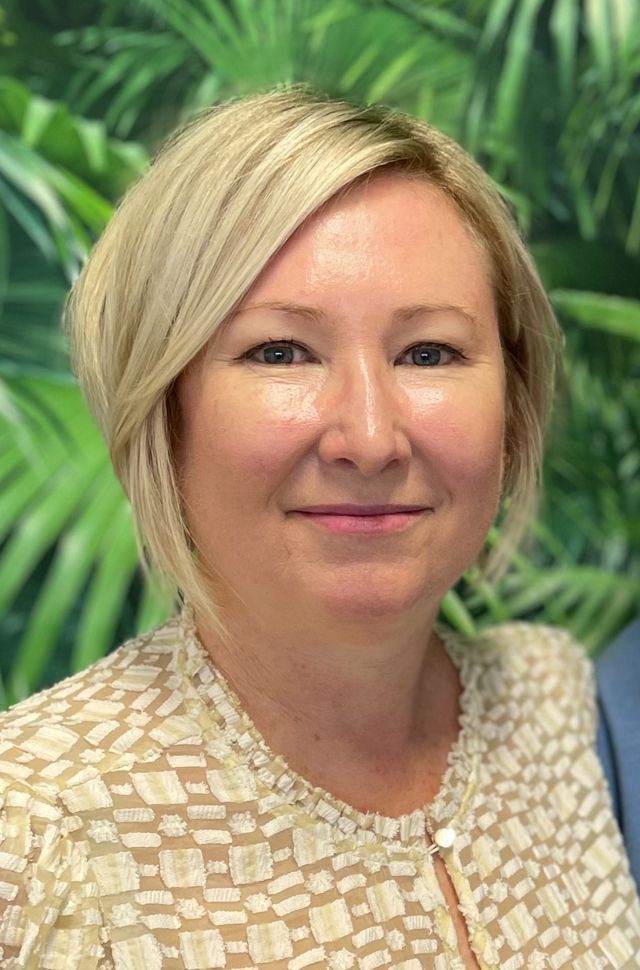 A smiling person with shoulder-length blonde hair, wearing a patterned white top in front of a green palm leaf background.