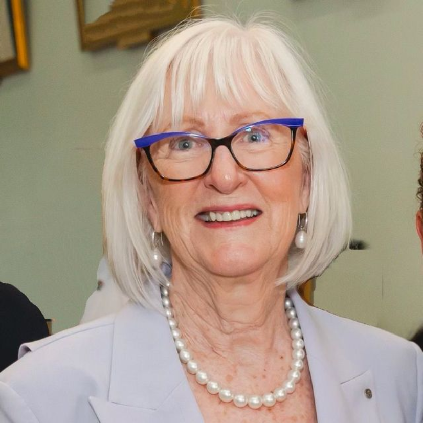 A person smiling, wearing a light blue blazer, a pearl necklace, and glasses with bold blue upper rims.