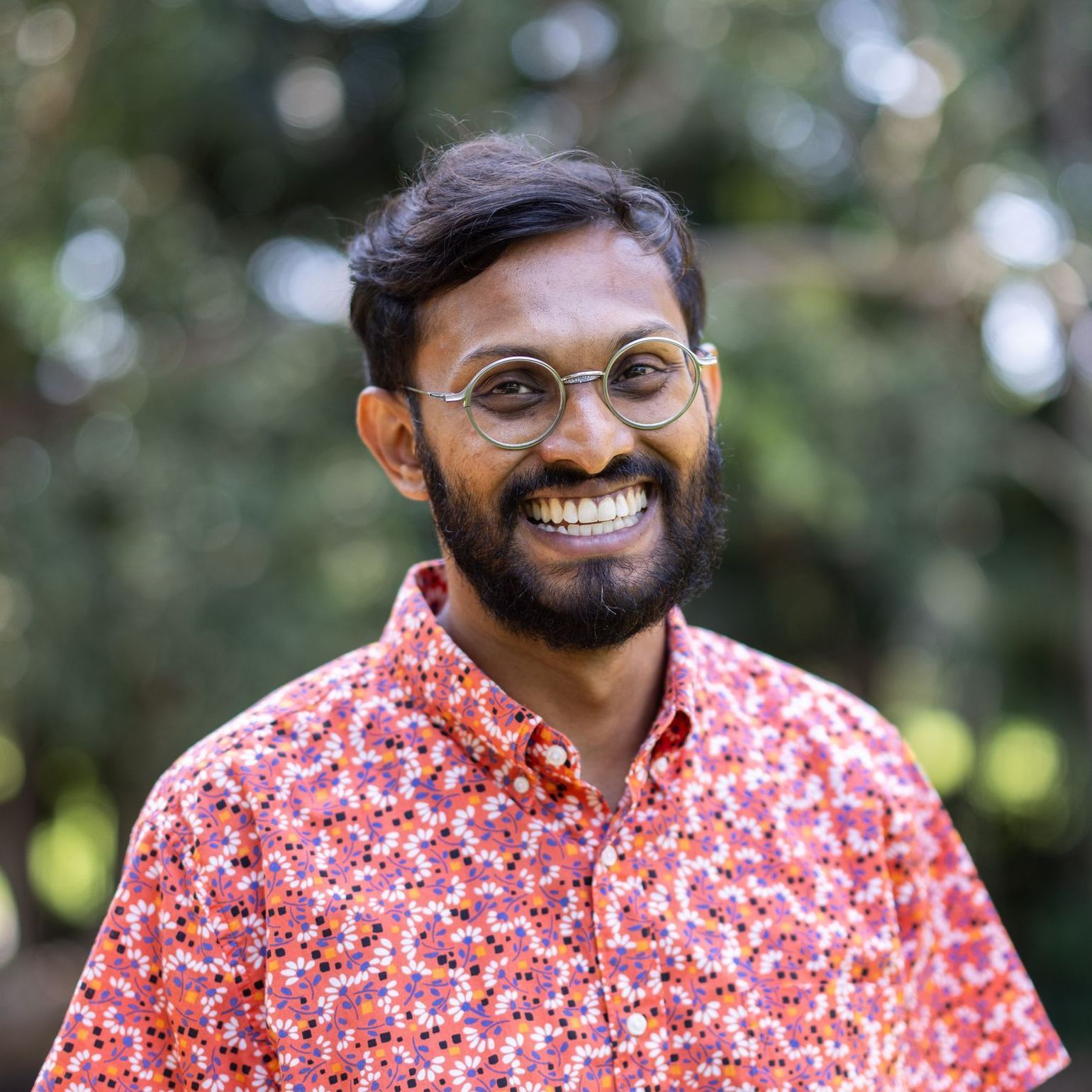 Smiling person with a beard and round glasses, wearing a pink floral shirt, outdoors against a blurred background.