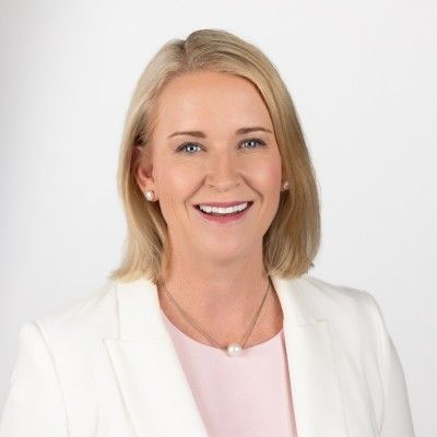 Professional headshot of Nicole Manison smiling, wearing a white blazer against a neutral grey background.